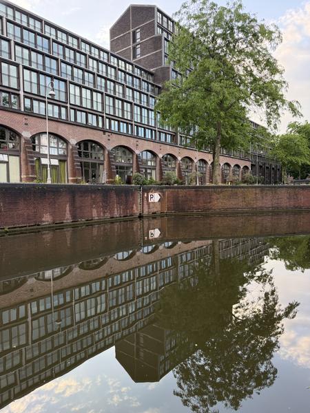A serene view captures a large brick building with numerous windows and arched ground-floor entrances, reflected perfectly in the calm waters of a canal at Plantage Kerklaan, Plantage, Amsterdam